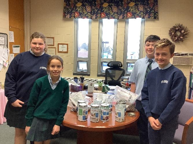 Four students from Saint Genevieve School, two boys and two girls, stand around a table. On the table are C4GC containers that will be used by each classroom to collect donations.